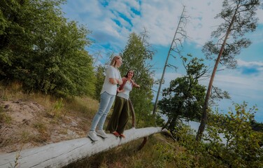 Naklejka premium Two women stand on a fallen tree trunk in a forest near a river, smiling and talking, enjoying nature, friendship, and outdoor adventure under a bright summer sky.