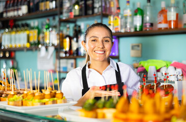Fototapeta premium Friendly young woman bartender offering appetizing pinchos in comfortable pub