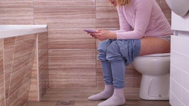 Woman sitting on toilet using smartphone in bathroom