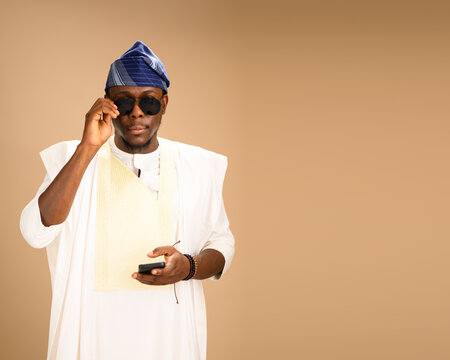 A Nigerian man dressed in agbada and blue fila adjusts his sunglasses while holding a phone, blending tradition and modern style in a studio portrait.