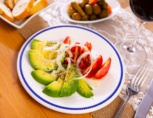 Vegetable salad of tomatoes, avocado and onions is complemented with bowl of green olives, small basket of bread, and red wine