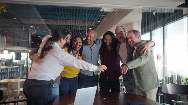 Multicultural colleagues gather in a tight huddle around a table, smiling and cheering after good news in a bright modern office, expressing unity, motivation and collaborative spirit.