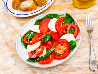 On plate are slices of mozzarella cheese and ripe tomatoes, decorated with basil leaves - traditional Italian salad caprese appetizer, served with carafe of olive oil and slices of baguette.