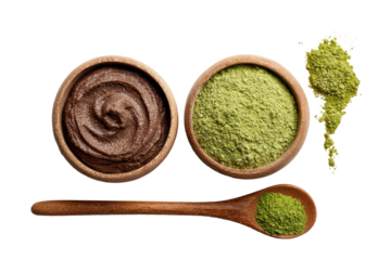 Two wooden bowls, one filled with dark brown paste, the other with light green powder, atop a light brown wooden spoon with more green powder.  A scattering of powder is above the green powder bowl