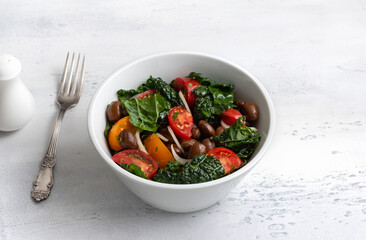 Fresh kale salad with beans, tomatoes and onion in white bowl on light background