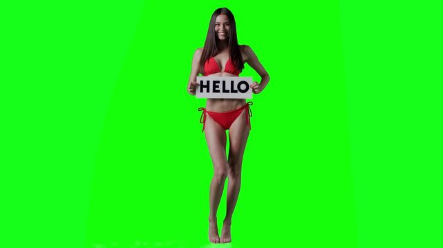 Real video of a smiling brunette woman in a red bikini holding a white sign with the word &ldquo;HELLO&rdquo; in bold black letters. The subject is isolated on a vivid green chroma key background, perfect for eas