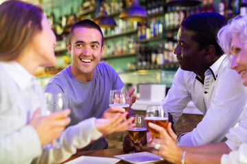 Company of multiracial friends drinking beer and talking with each other during the rest in a pub