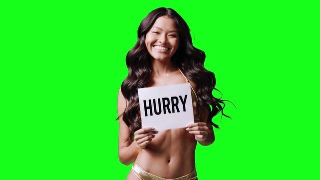 Real video of a cheerful brunette woman in a yellow bikini holding a white sign with the word &ldquo;HURRY&rdquo; in bold black letters. She stands isolated on a bright green chroma key background for easy compos
