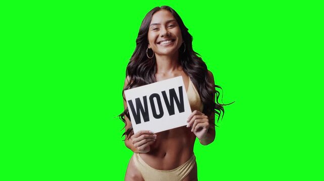 Real video of a smiling brunette woman in a golden bikini holding a white sign with the word &ldquo;WOW&rdquo; in bold black letters. The subject is isolated on a vivid green chroma key background, perfect for ea