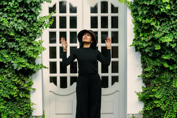 Stylish woman wearing black praying in front of ivy-covered wall