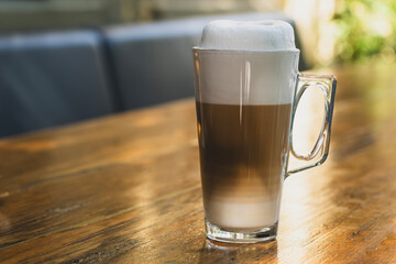 Foamy latte in a large glass on wooden table. Freshly made latte coffee served in tall glass. Large glass of latte with frothy milk on table. Hot latte with milk foam on rustic wooden surface.