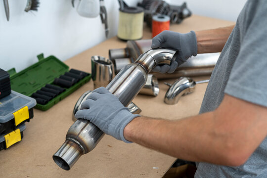 A person is handling a metal exhaust component while surrounded by various pipes and tools, preparing for installation in a workshop setting.