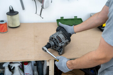 A person repairs a car alternator in a workshop, using a wrench while wearing gloves. The workbench showcases various tools and components for the repair.