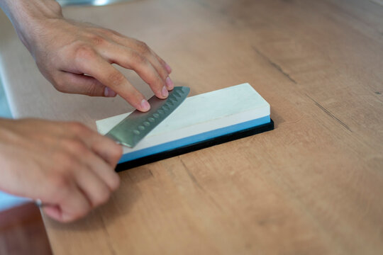A person is honing a kitchen knife against a whetstone on a wooden surface, focusing on sharpening the blade’s edge for better performance in food preparation.
