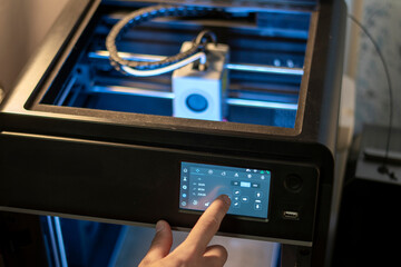 Hand is adjusting settings on a 3D printer with blue lighting in a home workspace. Focus is on the control interface as details are being fine-tuned.