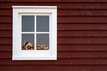 A dark red wooden exterior wall of a house with a four pane glass window. The antique building has wide white trim. The community of Bonavista, a church and house, is reflected in the mirrored glass