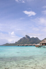 Overwater bungalows with Mount Otemanu views in the turquoise lagoon of Bora Bora, calm waters, and partly cloudy tropical sky in French Polynesia.