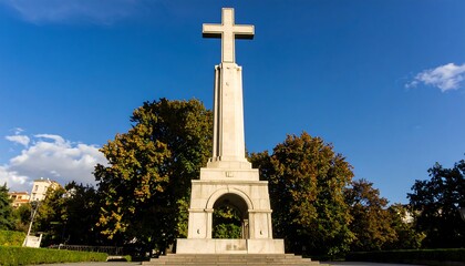 A tall, light-colored monument, featuring a large cross, stands prominently amidst lush parkland under a clear blue sky.