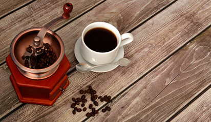 Manual coffee grinder, roasted beans and cup of hot black coffee with spoon on saucer on wooden table.