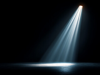 A single bright spotlight beams down onto a dark, empty floor, creating a focused, dramatic area of illumination amidst surrounding darkness