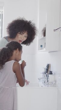 Vertical video: African American mother and daughter leaning at sink brushing teeth with toothbrush