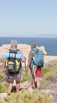 Vertical video: At dawn, senior couple hiking shore with poles, adjusting pack, glancing back
