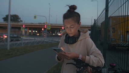 Young woman with neutral expression uses smartphone while wearing headphones and smartwatch near bicycle. Urban setting evokes serenity and contemplation.
