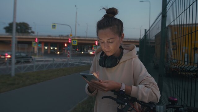 Young woman with neutral expression uses smartphone while wearing headphones and smartwatch near bicycle. Urban setting evokes serenity and contemplation. - Powered by Adobe