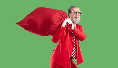 Modern, extravagant, young Santa Claus in red business suit screams with joy and happiness with huge, red bag filled with gifts on isolated green background. Christmas gift delivery.