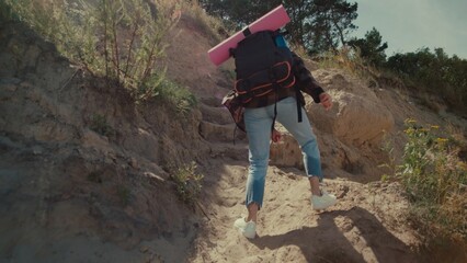 Determined young woman climbs hillside carrying backpack and yoga mat. Sunny day in nature evokes...