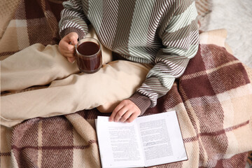 Beautiful young woman with cup of hot chocolate reading book on bed at home, closeup
