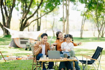 Joyful Asian family camping in a sunny garden, bonding over BBQ, tech, and laughter. Parents and daughter enjoy outdoor