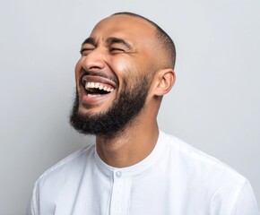 Obraz premium A man with a thick beard and a white collared shirt displays a joyous laugh against a neutral backdrop.
