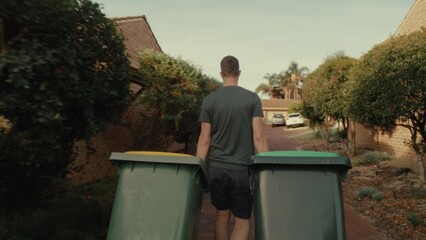 Young man carries two trash cans along suburban street. Serene atmosphere reflects daily routine in a residential neighborhood.