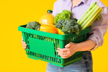 Woman holding green shopping basket with different fresh products on yellow background
