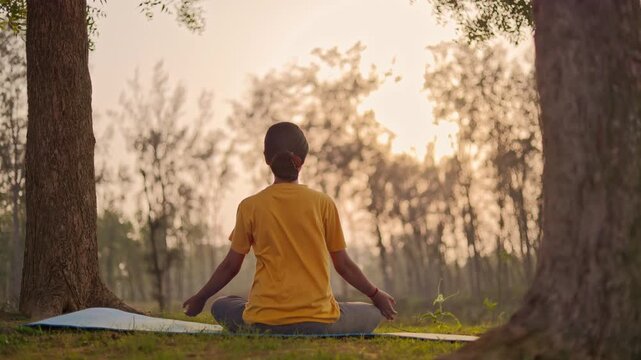 Back view of a woman sitting in padmasana pose practicing prayer pose in the forest during golden hour, maintaining healthy lifestyle.