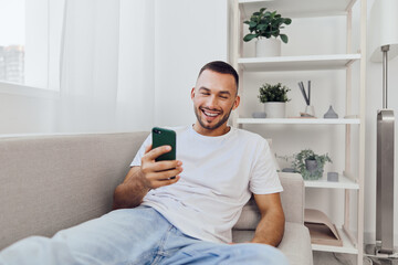 Happy man sitting on a couch, engaged with his smartphone in a cozy, modern living room, reflecting a relaxed lifestyle and connection to technology.