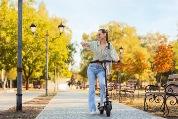 Fototapeta premium Beautiful young happy woman looking at wristwatch while riding electric kick scooter in park