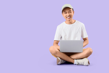 Young Asian man in cap using laptop on lilac background