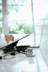 A doctor sits at a desk in a hospital office with stethoscope, documents, and laptop, providing healthcare consultation and care.