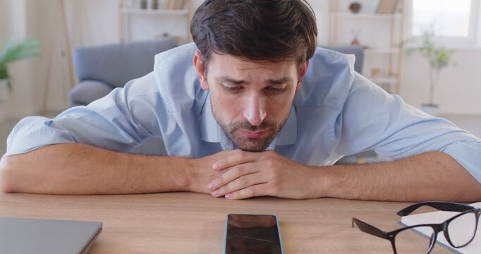 Nervous stressed young bearded man sitting at desk, looking at mobile phone screen, waiting impatiently for important call or message. Worried anxious guy leaning his head on table, feeling restless.