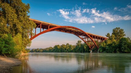 Fototapeta premium Long Rusty Orange Bridge Over Calm River Surrounded by Green Trees Under Blue Sky with Clouds