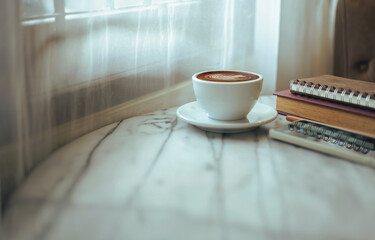 white cup of coffee with latte coffee and leisure reading books, diary on table by the window in cafe. vintage color tone background