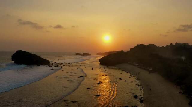 A stunning sunset over Watu Karung Beach in Pacitan, Indonesia, captures the golden hour light reflecting on the wet sand. Waves crash against rock formations, with the serene shoreline visible