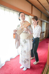 Obraz premium A woman in a kimono is being helped by another woman. The scene is set in a room with a red carpet and a window. Scene is one of assistance and support
