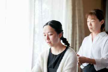 A woman is sitting in front of a window, looking out at the world. A woman is sitting behind her, braiding her hair. The two women are in a room with a window