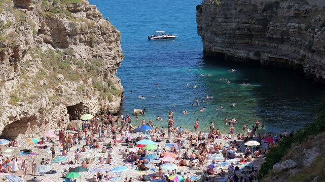 Sunny summer day in Polignano a Mare, Italy. Tourist boats float in the blue sea near crowded beaches and the historic stone bridge. Old buildings, cathedrals, and bustling streets. Mixed video
