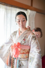 Obraz premium A woman in a kimono is adjusting her sash. The scene is set in a room with a window, and there are two other people in the background. The woman's outfit is traditional and elegant