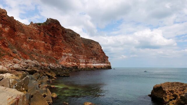 Smooth panoramic view of the vertical red cliffs and Bolata Beach near Balgarevo village, with calm sea and a blue canoe with two paddlers in the background.