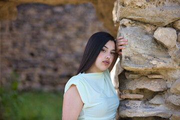 A beautiful Spanish teenager with long dark hair rests her hand on a stone wall and looks sad and serious. The young woman is alone, suffering from mental health issues and depression.
